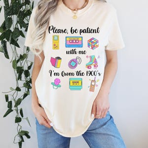 May include: A cream-coloured t-shirt featuring the phrase "Please, be patient with me, I'm from the 1900's" and various retro graphics. The design includes a cassette tape, Rubik's cube, roller skate, and a handheld game console.