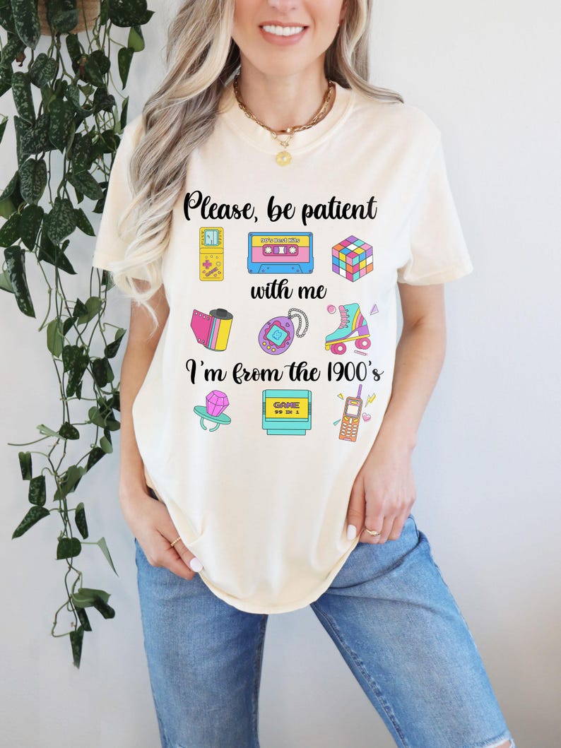 May include: A cream-coloured t-shirt featuring the phrase "Please, be patient with me, I'm from the 1900's" and various retro graphics. The design includes a cassette tape, Rubik's cube, roller skate, and a handheld game console.
