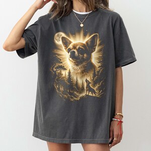 May include: Dark gray t-shirt featuring a detailed illustration of a Chihuahua with a radiant halo, set against a forest and moonlit sky. A smaller dog silhouette is howling. The design is in shades of brown and gold.