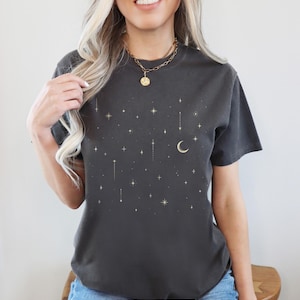 May include: Dark gray t-shirt with a celestial design featuring gold stars and a crescent moon. The shirt has a crew neck and short sleeves. The word "PEPPER" is printed at the bottom of the shirt.