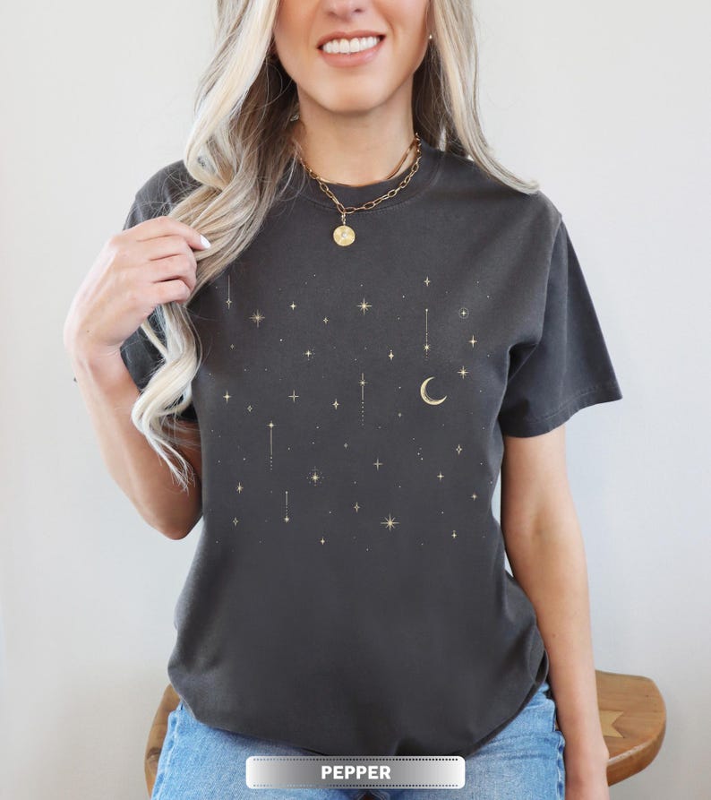 May include: Dark gray t-shirt with a celestial design featuring gold stars and a crescent moon. The shirt has a crew neck and short sleeves. The word "PEPPER" is printed at the bottom of the shirt.