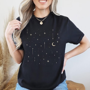 May include: Black t-shirt with a celestial design featuring gold stars, a crescent moon, and vertical lines. The word "BLACK" is displayed in a white rectangle at the bottom. The shirt is a crew neck style.