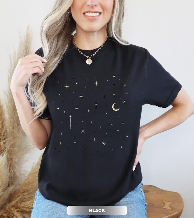 May include: Black t-shirt with a celestial design featuring gold stars, a crescent moon, and vertical lines. The word "BLACK" is displayed in a white rectangle at the bottom. The shirt is a crew neck style.