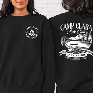 May include: Black sweatshirt with white text and graphic. The text reads "Camp Clara Lake Club" and "Est. 2024 Lake Tahoe Last Trail Before The Veil". The graphic is a campfire scene with trees and mountains.