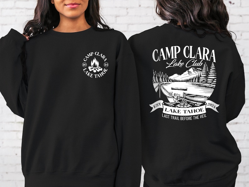 May include: Black sweatshirt with white text and graphic. The text reads "Camp Clara Lake Club" and "Est. 2024 Lake Tahoe Last Trail Before The Veil". The graphic is a campfire scene with trees and mountains.