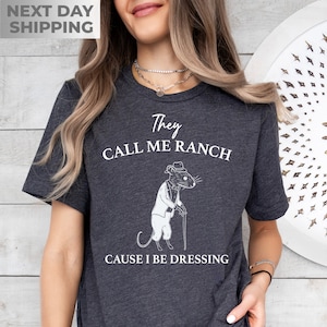 May include: A dark gray t-shirt with a white graphic of a rat wearing a tuxedo and holding a cane. The text on the shirt reads "They call me ranch cause I be dressing".