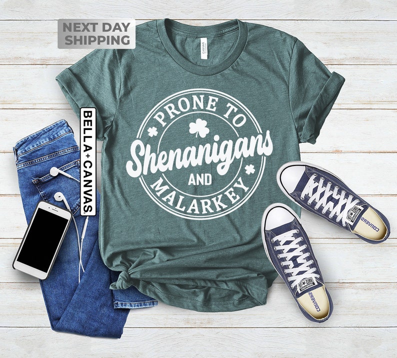 Op de afbeelding: Een tealgroen T-shirt met een witte cirkelvormige afbeelding met de tekst "*PRONE TO SHENANIGANS AND MALARKEY*" met een klavertje vier erin.