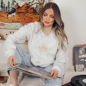 May include: A woman wearing a gray sweatshirt with a celestial design and blue jeans is holding a vinyl record. The record is on a turntable with a black record mat. There is a vintage tapestry on the wall behind her.