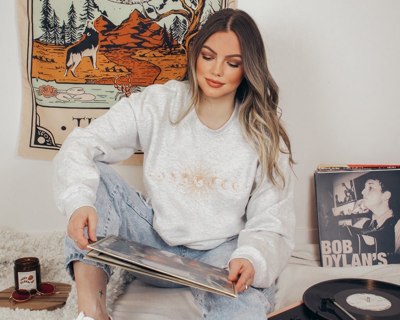 May include: A woman wearing a gray sweatshirt with a celestial design and blue jeans is holding a vinyl record. The record is on a turntable with a black record mat. There is a vintage tapestry on the wall behind her.