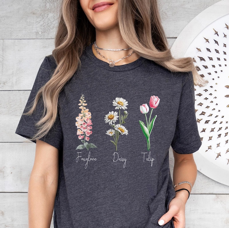 以下が含まれることがあります： ダークグレーのTシャツ。ジギタリス、デイジー、チューリップの花のイラストが描かれています。各画像の下に花の名前が書かれています。このTシャツはクルーネックで、半袖です。