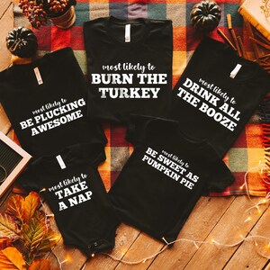 May include: Black t-shirts and a onesie with white text. The shirts say "most likely to BURN THE TURKEY", "most likely to DRINK ALL THE BOOZE", "most likely to BE PLUCKING AWESOME", and "most likely to BE SWEET AS PUMPKIN PIE". The onesie says "most likely to TAKE A NAP".