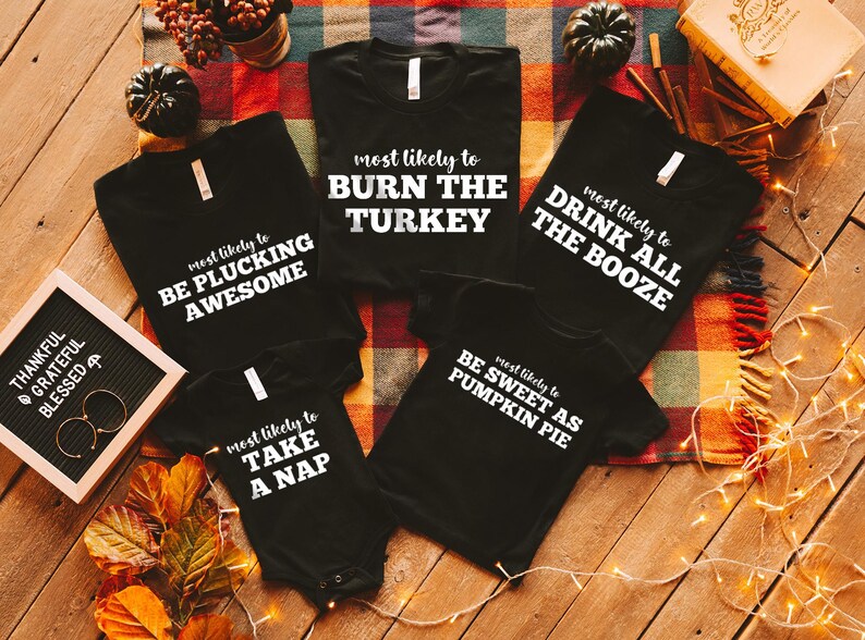 May include: Black t-shirts and a onesie with white text. The shirts say "most likely to BURN THE TURKEY", "most likely to DRINK ALL THE BOOZE", "most likely to BE PLUCKING AWESOME", and "most likely to BE SWEET AS PUMPKIN PIE". The onesie says "most likely to TAKE A NAP".