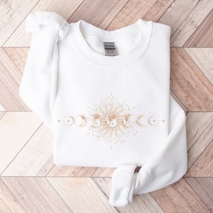 May include: White sweatshirt with a gold sun and moon design. The design features a sun with rays and a crescent moon on either side.