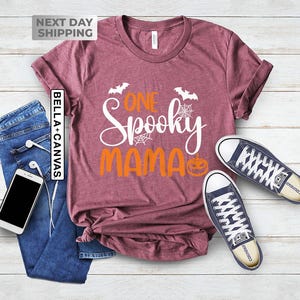 May include: A maroon Bella+Canvas t-shirt with the text "One Spooky Mama" in white and orange. The design includes bats, spiderwebs, and a jack-o'-lantern. The shirt is paired with blue jeans and a pair of navy blue sneakers.
