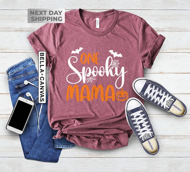 May include: A maroon Bella+Canvas t-shirt with the text "One Spooky Mama" in white and orange. The design includes bats, spiderwebs, and a jack-o'-lantern. The shirt is paired with blue jeans and a pair of navy blue sneakers.