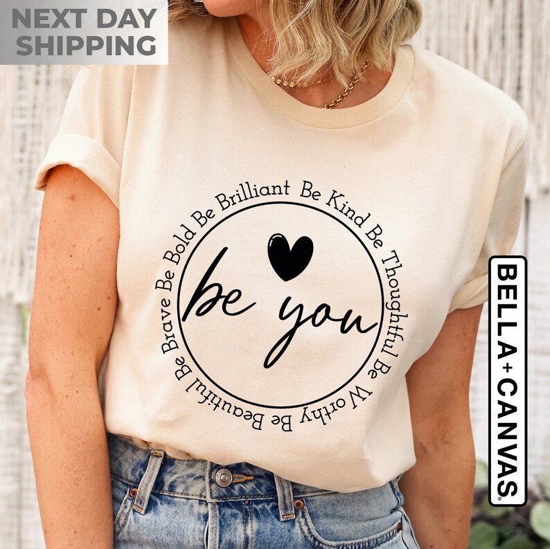 May include: A beige t-shirt with a black circle design that says "be you" with a heart in the center. The circle is surrounded by the words "Be Kind Be Thoughtful Be Worthy Be Beautiful Be Brave Be Bold Be Brilliant".