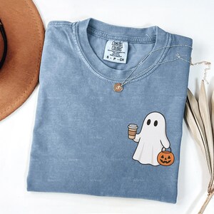 May include: A blue jean coloured t-shirt featuring a ghost graphic holding a coffee cup and a jack-o'-lantern. The shirt has a crew neck and short sleeves. A silver necklace with a pendant is on the shirt. The shirt is labelled "Comfort Colors".