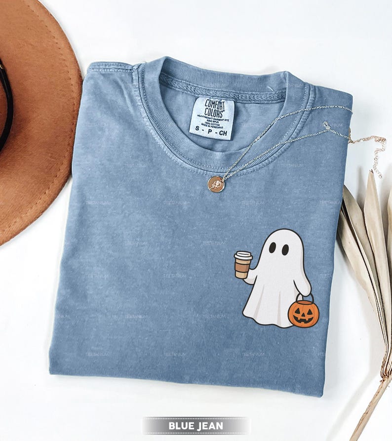 May include: A blue jean coloured t-shirt featuring a ghost graphic holding a coffee cup and a jack-o'-lantern. The shirt has a crew neck and short sleeves. A silver necklace with a pendant is on the shirt. The shirt is labelled "Comfort Colors".