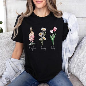 以下が含まれることがあります： 黒いTシャツ。フォックスグローブ、デイジー、チューリップの水彩イラストが描かれています。花の名前が下に書かれています。Tシャツはクルーネックです。