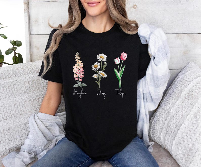 以下が含まれることがあります： 黒いTシャツ。フォックスグローブ、デイジー、チューリップの水彩イラストが描かれています。花の名前が下に書かれています。Tシャツはクルーネックです。
