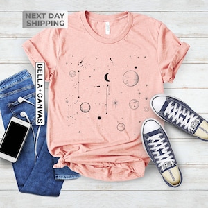 May include: A pink t-shirt with a black graphic of a crescent moon, stars, and planets. The shirt is laid flat on a wooden surface with a pair of blue converse shoes.