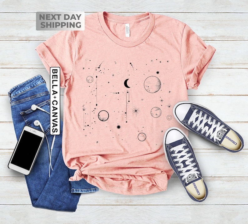 May include: A pink t-shirt with a black graphic of a crescent moon, stars, and planets. The shirt is laid flat on a wooden surface with a pair of blue converse shoes.