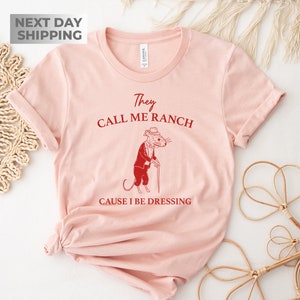 May include: Pink t-shirt with a red graphic of a mouse wearing a hat and holding a cane. The text on the shirt reads "They Call Me Ranch Cause I Be Dressing".