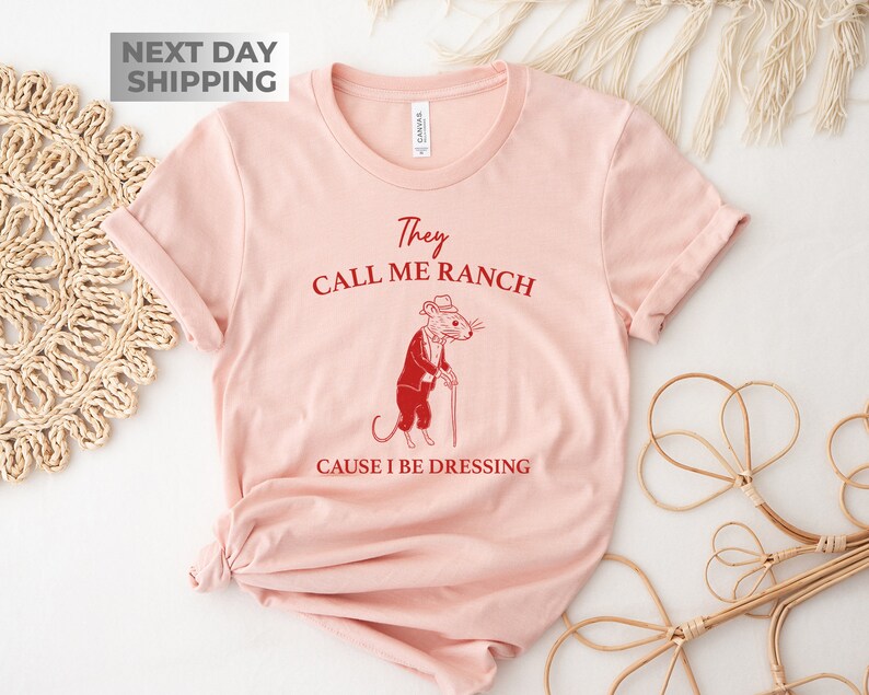 May include: Pink t-shirt with a red graphic of a mouse wearing a hat and holding a cane. The text on the shirt reads "They Call Me Ranch Cause I Be Dressing".