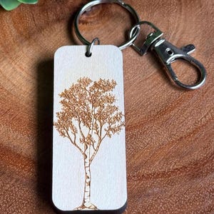 May include: A rectangular wooden keychain with a detailed tree design. The tree is etched in brown against a light background. The keychain has a silver metal ring and clasp for attaching to keys or bags.