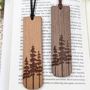 May include: Two wooden bookmarks featuring a forest tree design. One is light brown, the other dark brown. Each has a leather cord at the top. The bookmarks are rectangular with a rounded base. Ideal for book lovers.