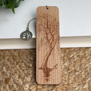 May include: Wooden bookmark with a detailed engraving of a weeping willow tree and a small bird. The bookmark is attached to a silver-colored chain with a tree-shaped charm. The wood has a natural grain and a warm, light brown color.