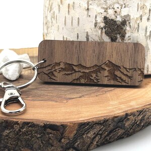 May include: A rectangular wooden keyring with a mountain range design. The keyring is made of dark brown wood and features a detailed engraving of a mountain landscape. It includes a silver metal keyring and clasp.
