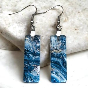 May include: A pair of rectangular dangle earrings with a marbled blue and white design. The earrings have silver-coloured hooks and connectors. The marbled pattern features swirls of dark and light blue with hints of gold and white, creating a unique, artistic look.