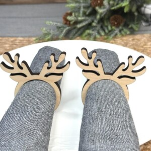 May include: Two wooden reindeer antler napkin rings on grey napkins. The napkin rings are light brown with a cutout antler design. The napkins are rolled up and placed on a white plate. Decorative greenery and pinecones are in the background.