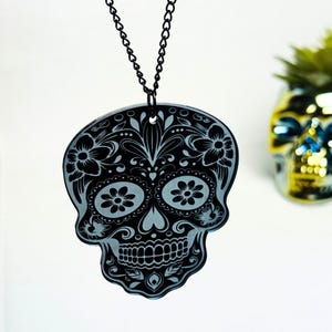May include: A black skull pendant with floral designs and a black chain. The pendant is a stylized representation of a skull, with intricate floral patterns filling the eye sockets and surrounding the skull's features. The pendant is hanging against a white background.