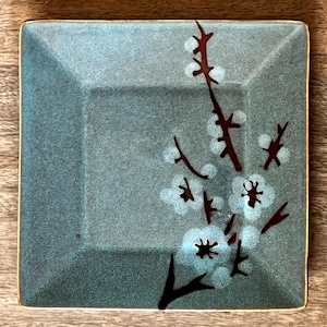 May include: Square, teal-colored ceramic dish with a gold rim. The dish features a floral design of white blossoms and brown branches. The dish is placed on a wooden surface.