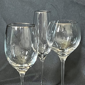 May include: Three clear wine glasses with gold rims are displayed against a dark background. The glasses include a champagne flute, a standard wine glass, and a larger, round wine glass. The stems and bases are clear.