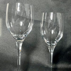 May include: Two clear, stemmed wine glasses. The glasses have a classic design with a rounded bowl and a long, slender stem. The rims of the glasses have a decorative, textured pattern. The glasses are made of clear glass and are set against a dark background.