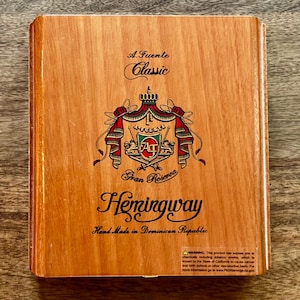 May include: Wooden cigar box with the text "A. Fuente Classic" and "Hemingway" in elegant script. The box features a detailed emblem with red and gold accents, and the words "Gran Reserva" are also visible.