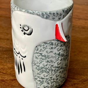 May include: A ceramic owl-themed tumbler. The tumbler features a white owl face with black details and a red beak. The body of the tumbler is speckled gray and white. The rim is gray. The tumbler is on a wooden surface.