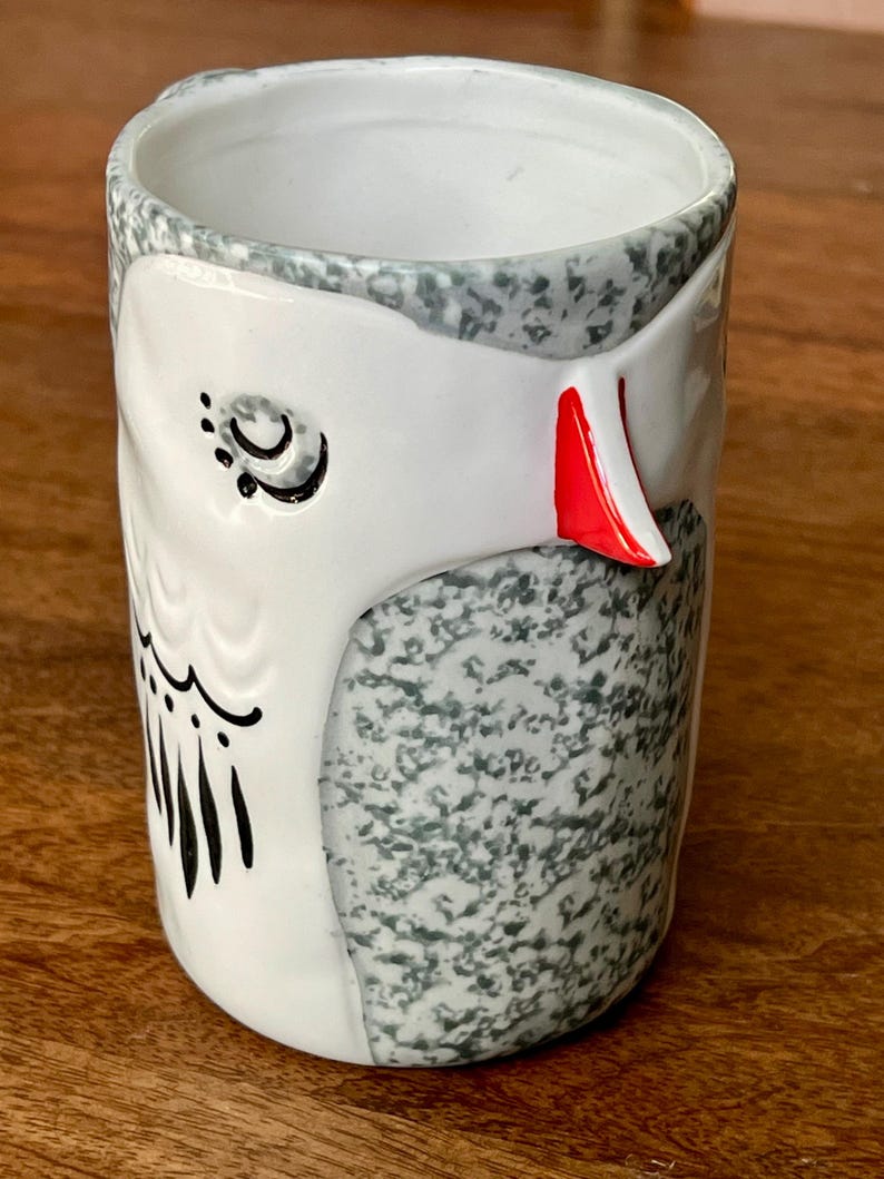 May include: A ceramic owl-themed tumbler. The tumbler features a white owl face with black details and a red beak. The body of the tumbler is speckled gray and white. The rim is gray. The tumbler is on a wooden surface.