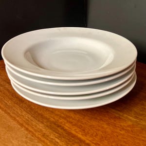 May include: A stack of four white, shallow soup bowls. The bowls have a simple, classic design with a slightly raised rim. They are stacked neatly on a wooden surface, with a dark background.