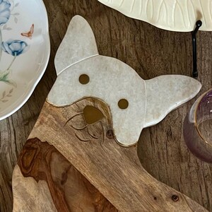 Op de afbeelding: Een houten serveerplank in de vorm van een hondenkop met een witte marmeren inleg voor het gezicht. De plank heeft twee gouden accenten voor de ogen en een gegraveerde neus.
