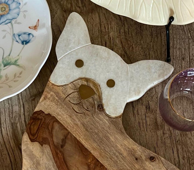 Op de afbeelding: Een houten serveerplank in de vorm van een hondenkop met een witte marmeren inleg voor het gezicht. De plank heeft twee gouden accenten voor de ogen en een gegraveerde neus.