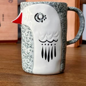 May include: Ceramic mug with a bird design. The mug features a white bird face with a red beak, black eye details, and black feather accents. The mug's body has a speckled gray and white pattern, with a curved handle.