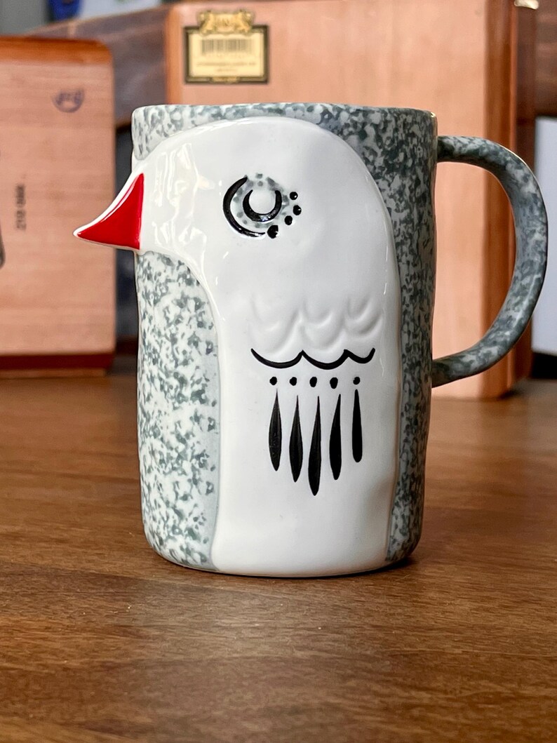 May include: Ceramic mug with a bird design. The mug features a white bird face with a red beak, black eye details, and black feather accents. The mug's body has a speckled gray and white pattern, with a curved handle.