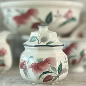May include: A white ceramic sugar bowl with a lid, featuring a floral design with pink and green flowers. The bowl has a hexagonal shape and a blue trim around the top.