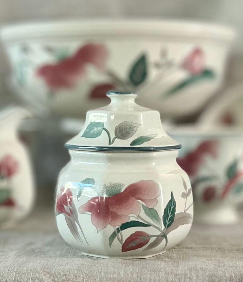 May include: A white ceramic sugar bowl with a lid, featuring a floral design with pink and green flowers. The bowl has a hexagonal shape and a blue trim around the top.