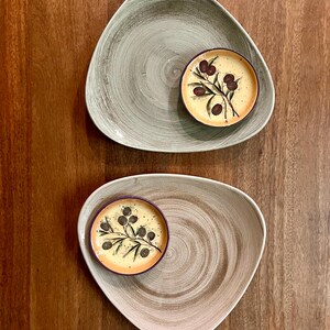 May include: Two triangular ceramic plates with a textured, swirled finish in sage green and light brown. Each plate features a small, round ceramic dish with a cream-colored center, a purple rim, and a painted olive branch illustration.