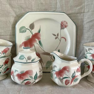 Puede incluir: Un juego de tazas de té y platillos con estampado floral, una jarra de crema, un azucarero y un plato para servir. El estampado floral presenta flores rosas y hojas verdes sobre un fondo blanco.